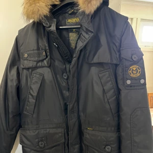 Svart vinterjacka från Wiggy's med Äkta päls  - Vinter jacka från Wiggy's i svart nylon med varm Äkta päls runt huvan. Jackan har flera fickor med knappar, patch på ärmen och dragkedja samt knäppning framtill. Fodrad med polyester och lamalite-isolering för extra värme. Perfekt för kalla dagar.