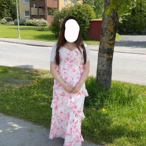 Vit långklänning med rosa blommor - Använd en gång! Säljer en romantisk vit långklänning med rosa blommigt mönster och volangdetaljer. Klänningen har v-ringad hals och är ärmlös, perfekt för varma dagar. Lätt och luftig design som ger en somrig känsla.