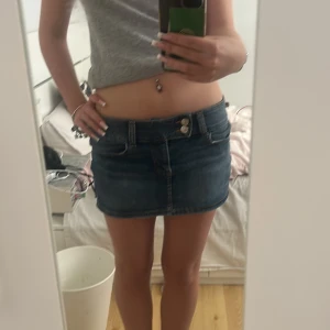 Blå jeanskjol med fickor - Säljer en klassisk blå jeanskjol i kort modell med två knappar framtill, bälteshällor och stora bakfickor. Kjolen har en enkel och tidlös design som är lätt att matcha med olika toppar. Perfekt för dig som gillar denim och vill ha en snygg bas i garderoben.