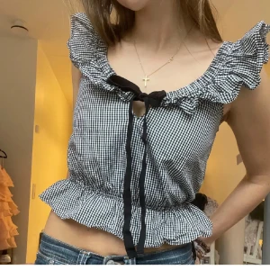 Rutig volangblus från Zara  - Supersöt svartvit rutig blus från Zara med volangkant vid axlar och nederdel. Blusen är croppad, har knytband framtill och är ärmlös. Perfekt för dig som gillar romantiska detaljer och en trendig look. Storlek XS passar också S