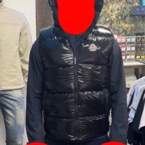 Svart glansig dunväst från Moncler - Svart glansig dunväst från Moncler med huva och dragkedja framtill. Klassisk Moncler-logga på bröstet och quiltad design. Perfekt för lager-på-lager och ger en clean streetstyle-vibe.