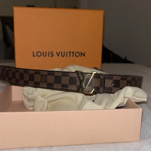 Brunt Louis Vuitton bälte i skinn till dam - Säljer ett brunt Louis Vuitton bälte i skinn med klassiskt rutigt mönster och en stor LV-logga i guld som spänne. Bältet har en lyxig känsla och passar perfekt till jeans eller kostymbyxor. Kommer med originalbox och dustbag.