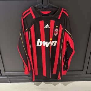 AC Milan långärmad Kaka' 22 tröja - Säljer en klassisk AC Milan långärmad fotbollströja från Adidas med Kaka' och nummer 22 på ryggen. Tröjan är svart och röd med vertikala ränder, guldiga detaljer och klubbmärke på bröstet. Materialet är lätt och andas, perfekt för fotbollsälskare.