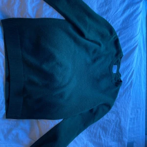 Mörkgrön merinoullströja Colorful Standard - Säljer en stilren mörkgrön stickad tröja från Colorful Standard i 100% merinoull. Tröjan har rund halsringning, ribbade muddar och lång ärm. Perfekt för dig som gillar klassisk och enkel stil med premiumkänsla.