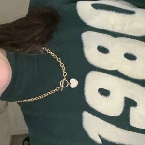 Guldfärgat halsband med hjärthänge - Snyggt guldfärgat halsband med grov kedja och ett sött vitt hjärthänge. Halsbandet har en trendig toggle-låsning framtill som ger en cool detalj. Perfekt accessoar för att lyfta din outfit med en touch av guld och kärlek.