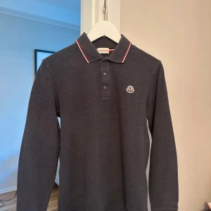 Moncler Piké Långärmad - Snygg mörkgrå långärmad pikétröja från Moncler med klassisk krage och knäppning framtill. Kragen har röda och vita ränder. Bröstet pryds av Monclers ikoniska logga. Perfekt för dig som gillar stilrena och exklusiva plagg. Tveka inte att höra av dig vid frågor eller fler bilder!