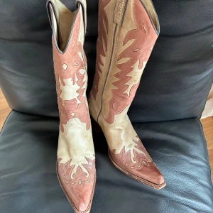 Sancho cowboy boots  - Säljer ett par handgjorda Sancho cowboy boots i beige och brun skinn med snygga broderade detaljer och spetsig tå. Boots med hög klack och klassiskt westernmönster, tillverkade i Spanien. Perfekta för dig som vill sticka ut med en unik stil.