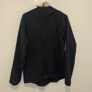 Svart vindjacka från Nike med huva - Svart vindjacka från Nike med minimalistisk design och broderad swoosh på bröstet. Jackan har hel dragkedja, justerbar huva och två sidofickor med dragkedja. Perfekt för blåsiga dagar och enkel att matcha med allt.