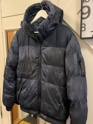 Mörkblå pufferjacka från Etrel XS - Säljer en mörkblå pufferjacka från Etrel i storlek XS. Jackan har huva med snörning, dragkedja framtill och fickor på sidorna. Materialet är syntet och jackan har ett diskret mönster som ger en cool vibe. Perfekt för kalla dagar och enkel att matcha med din stil.