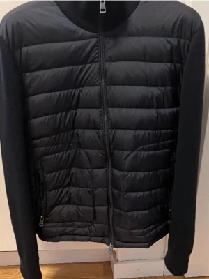 Svart dunjacka från Moncler - Svart dunjacka från Moncler med quiltad framsida och stickade ärmar. Jackan har dragkedja framtill, två sidofickor med dragkedja och Moncler-logga på ärmen. Perfekt för dig som vill ha en stilren och varm jacka med snygg passform. Kvitto finns !! Pris kan diskuteras vid bra affär. Skriv för mer bilder
