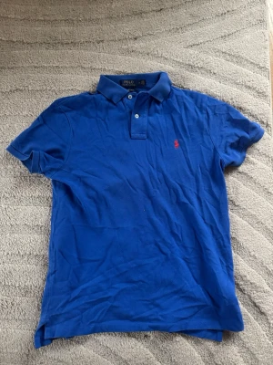 Blå piké från Polo Ralph Lauren - Säljer en klassisk blå pikétröja från Polo Ralph Lauren i custom slim fit. Pikén har korta ärmar, krage och två knappar framtill. Den ikoniska röda logon är broderad på bröstet. Perfekt för dig som gillar stilrena och sportiga plagg.