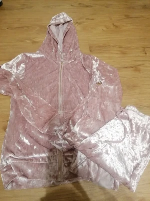 Rosa mjukisbyxor & hoodie i sammet - Säljer ett par mjukisbyxor & hoodie i ljusrosa sammet med glansig finish. Byxorna har resår i midjan med snörning och muddar vid bensluten. Perfekta för chill eller när du vill vara extra comfy. Matchande hoodie ingår också. 