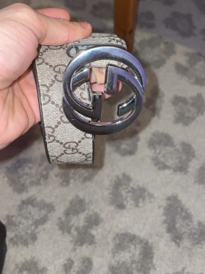 Gucci beige skärp med GG-spänne - Säljer ett snyggt Gucci-skärp i beige med klassiskt GG-mönster och ett stort, silverfärgat GG-spänne. Skärpet är gjort i skinn och har detaljer i brunt på insidan. Perfekt för att lyfta din outfit med en lyxig touch.