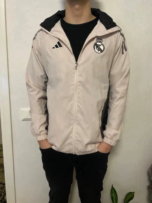 Beige vindjacka/windbreaker Real Madrid Adidas - Säljer en beige vindjacka från Adidas X Real Madrid. Jackan har svarta detaljer på axlarna och dragkedja framtill. Perfekt för dig som gillar sportig stil och vill visa ditt lag. Den har använts några gånger och har inga defekter. Den är storlek M,  ställ frågor vid minsta fundering 😊✨ 