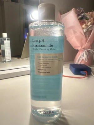 COSRX Low pH Niacinamide Micellar Cleansing Water - Micellär rengöringsvatten med låg pH och niacinamid, perfekt för känslig hud. Innehåller zink PCA, allantoin och hyaluronsyra för att ta bort smink, kontrollera sebum och återfukta. Kommer i en genomskinlig plastflaska med blått vatten, 400 ml.