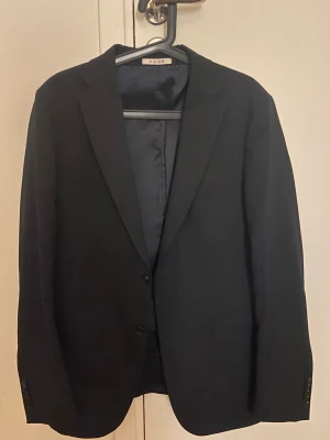 Mörkblå Kavaj - Stilren svart kava med klassisk passform och två knappar framtill. Kavajen har slät yta, diskreta fickor och helfodrad insida. Perfekt för dig som vill ha en enkel och snygg look till vardagen eller speciella tillfällen.