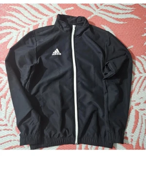 Svart vindjacka från Adidas - Svart vindjacka från Adidas med vit dragkedja och logga på bröstet. Jackan har hög krage, långa ärmar och elastiska muddar vid ärmslut och nederkant. Perfekt för träning eller chill dagar. Materialet är lätt och känns sportigt.