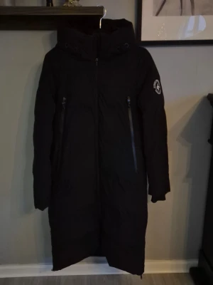 Svart lång dunjacka från Cross Sportswear - Säljer en svart lång dunjacka från Cross Sportswear med huva och två stora dragkedjefickor framtill. Stängs med dragkedja hela vägen upp och har en stilren look. 