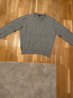 Grå v-ringad tröja Tommy Hilfiger XL - Snygg grå v-ringad tröja från Tommy Hilfiger i premium bomull. Klassisk design med diskret logga på bröstet och ribbade muddar vid ärmslut och nederkant. Perfekt för dig som gillar stilrena och bekväma plagg.