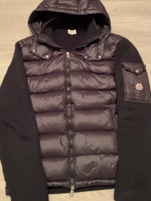 Svart Moncler pufferjacka  - Svart pufferjacka från Moncler med stickade ärmar och huva. Jackan är i helt nytt skick där alla orginal taggar följer med. Jackan fungerar som den ska och märket scannar som den ska. Jackan är i Moncler storlek xl som då är L i vanliga storlekar, skriv gärna för mer information 