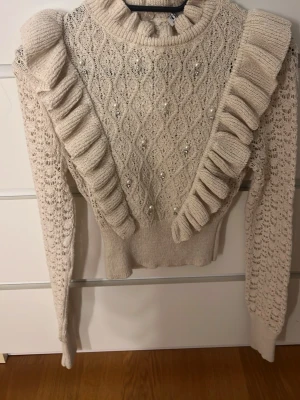 Beige stickad tröja med volanger Zara - Superfin beige kort stickad tröja från Zara med volangdetaljer längs axlarna och pärlor på framsidan. Tröjan har ett snyggt spetsmönster, ribbad krage och långa ärmar. 