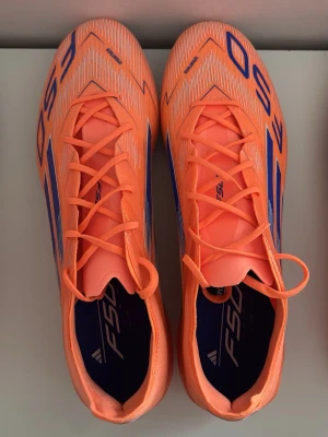 Adidas F50 CORAL BLAZE  - Säljer ett par Adidas F50 fotbollsskor i en riktigt fet orange färg med blå detaljer och vita linjer. Skorna har snörning och platt sula med dobbar för gräsplan. Lätta och snygga med en sportig vibe, perfekta för dig som vill sticka ut på planen.