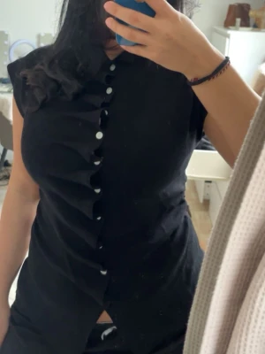 Svart blus med volang och knappar - Säljer en svart blus med korta ärmar och snygg volang längs knappraden framtill. Blusen har en klassisk krage och vita knappar som kontrast mot det svarta tyget. Perfekt för dig som gillar stilrena detaljer och vill ha något som sticker ut.aldrig använd 