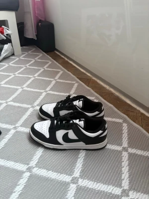 Nike Dunk Low( Pandas) - Nike Dunk Low sneakers i svart och vitt med klassisk swoosh på sidan. Skorna har snörning, perforerad tå och platt sula. Tillverkade i syntet och skinn för en clean och sportig look. Perfekt för dig som gillar streetwear och vill ha en ikonisk sneaker i garderoben.