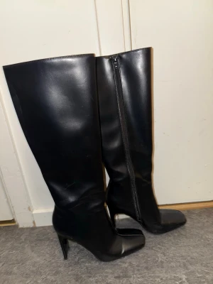 Svarta höga stövlar med klack - Säljer ett par svarta höga stövlar med spetsig tå och markerad klack. Stövlarna har dragkedja på insidan och är gjorda i slätt skinnliknande material. Snygg och stilren design .