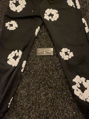 Svarta Denim Tears byxor med vit print - Svarta byxor från Denim Tears med stora vita blommönster tryckta över hela plagget. Byxorna har en avslappnad passform och resår vid bensluten. Materialet är bomull och designen är riktigt unik och sticker ut. Perfekt för dig som vill ha något som syns.