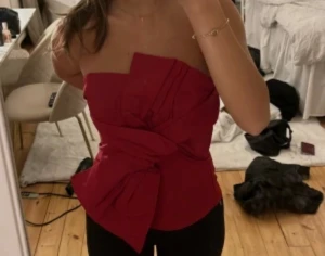 Röd axelbandslös topp med rosett - Säljer en snygg röd axelbandslös topp med stor rosettdetalj framtill. Toppen har en croppad passform och är tillverkad i ett glansigt material som ger en lyxig känsla. Perfekt för dig som vill sticka ut och ha något unikt på överkroppen. Använd 1 gång 