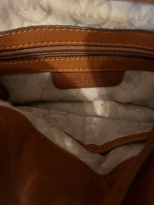 Brun handväska från Michael Kors - Säljer en snygg brun handväska från Michael Kors i skinn med guldfärgade metalldetaljer och kedja på axelremmen. Väskan har ett klassiskt MK-mönstrat innerfoder, dragkedja och praktisk nyckelhållare. Perfekt storlek för allt du behöver ha med dig.