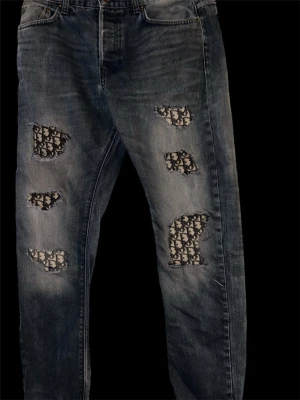 Blå jeans med Di0R patchar - Unika blå jeans med flera patchar i Di0r -mönster på framsidan. Jeansen har en klassisk femficksdesign och är tillverkade i slitstarkt bomullsmaterial. Passformen är rak och de har en cool, vintage tvätt med tydliga slitningar och broderade detaljer. Bilden visar hur jeansen sitter 
