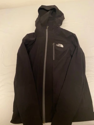 Svart vindjacka från The North Face - Svart vindjacka från The North Face med huva och hel dragkedja framtill. Jackan har flera praktiska fickor med dragkedja och meshpaneler vid axlarna för extra ventilation. Perfekt för dig som gillar att vara aktiv utomhus och vill ha en stilren look.