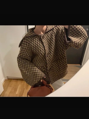 Brun rutig kappa med stora knappar - Säljer en brun och beige rutig kappa med bred krage och stora knappar framtill. Jackan har en loose passform och är tillverkad i ett ulligt material som känns mjukt och varmt. Endast använd fåtal gånger så i väldigt bra skick!