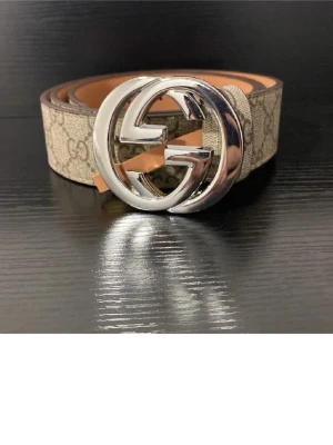  beige bälte - Säljer ett bälte i beige med klassiskt monogrammönster och stor silverfärgat spänne. Bältet är gjort i skinn och canvas, med snygga detaljer och lyxig känsla. Perfekt för att lyfta din outfit.