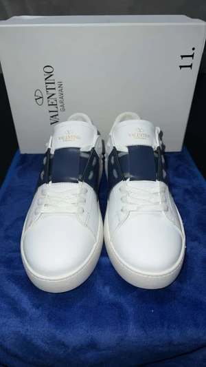 Valentino Garavani Open sneakers vit/blå - Snygga Valentino Garavani Open sneakers i vitt skinn med bred marinblå läderdetalj över snörningen. Klassisk rund tå, platt sula och diskret guldlogga på plösen. Perfekta för dig som vill ha en clean och lyxig look med en twist.