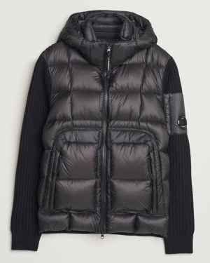 Svart dunjacka från C.P. Company - Säljer en svart dunjacka från C.P. Company med stickade ärmar och huva. Jackan har quiltad front, två stora fickor med dragkedja och en ikonisk linsdetalj på ärmen. Materialmix av dun och stickad polyester ger en unik look.