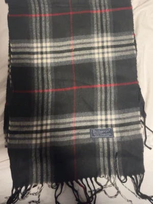 Rutig scarf från Burberry - Säljer en klassisk rutig scarf från Burberry i 100% lammull. Scarfen har svart, vit och grå bas med röda detaljer och fransar nedtill. Perfekt för dig som vill ha en tidlös och snygg look med brittisk känsla.