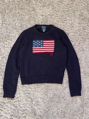 Mörkblå stickad tröja Polo Ralph Lauren - Snygg mörkblå stickad tröja från Polo Ralph Lauren i storlek S. Framsidan har en stor amerikansk flagga i rött, vitt och blått samt RL-broderi. Tröjan har rund hals, ribbade muddar och är långärmad. Perfekt för dig som gillar klassisk och preppy stil.