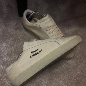 Saint Laurent vita sneakers - Säljer ett par stilrena vita sneakers från Saint Laurent med handskriven logga på sidan och guldfärgad text på plösen. Skorna har rund tå, snörning och platt sula. Materialet är en mix av skinn och canvas, vilket ger en cool och lyxig känsla. Har tyvärr slängt boxen, därav priset