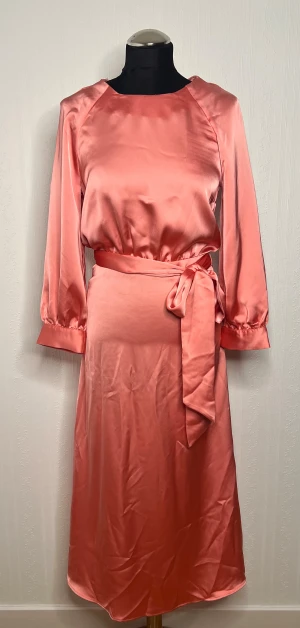 Ljusrosa långklänning i satin  - Säljer en elegant ljusrosa långklänning från H&M i glansigt satinmaterial. Klänningen har långa ärmar med lätt puff och ett brett knytband i midjan som ger en snygg siluett. Perfekt för dig som gillar stilrena och feminina plagg.