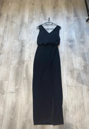 Svart glittrig långklänning från Vila - Svart långklänning från Vila med glittrigt tyg och omlott upptill. Klänningen har v-ringad hals och är ärmlös, vilket ger en elegant look. Materialet är syntet med en glansig finish på insidan. Perfekt för dig som vill sticka ut med stil.