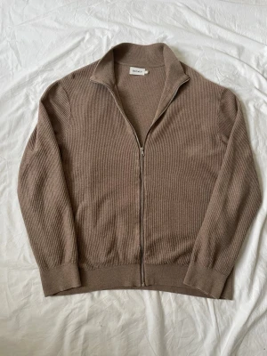 Beige ribbad kofta från East West | L  - Säljer en beige ribbad kofta från East West i storlek XL. Koftan har hel dragkedja framtill, hög krage och långa ärmar. Materialet är stickat och ger en mjuk och skön känsla. Perfekt för lager-på-lager under kyliga dagar.