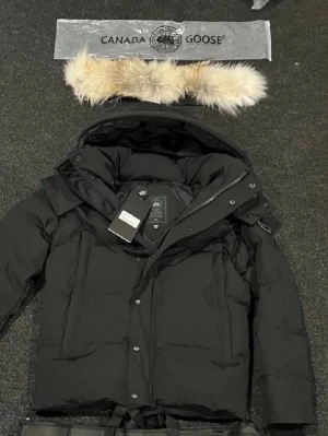 Svart dunjacka från Canada Goose - Svart dunjacka från Canada Goose med avtagbar huva och beige pälsdetalj. Jackan har dragkedja och knappar framtill, samt en snygg patch på ärmen. Perfekt för kalla vinterdagar och har en clean, stilren look.