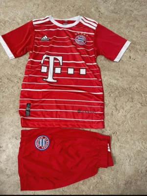 FC Bayern München fotbollströja & shorts - Säljer ett rött FC Bayern München matchställ med tröja och shorts. Tröjan har vita ränder, klubbmärke, sponsortryck och nummer 10 med Sané på ryggen. Shorts med klubbmärke. Perfekt för fotbollsfans som vill ha en snygg och bekväm outfit på planen.