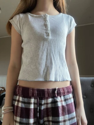 Ljusgrå ribbad topp med knappar - Säljer en ljusgrå ribbad topp med korta ärmar och rund hals. Toppen har dekorativa knappar framtill och en liten spetskant längs knappraden. Croppad passform som ger en chill vibe, perfekt till jeans eller shorts.