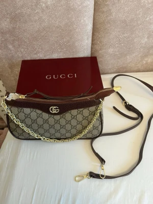 Gucci brun axelväska med kedja - Säljer en snygg axelväska från Gucci med klassiskt GG-mönster i beige och brunt. Väskan har guldfärgad kedja, bruna skinn-detaljer och dragkedja upptill. Perfekt storlek för det viktigaste och har en lyxig vibe med ikonisk logga framtill.