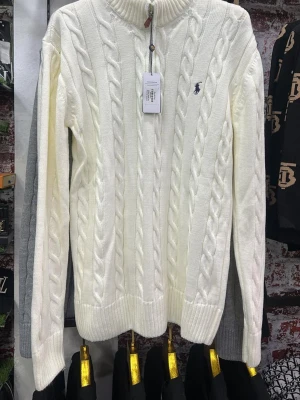 Vit polotröja från Ralph Lauren - Säljer en vit polotröja från Ralph Lauren med klassiskt flätstickat mönster och broderad logga på bröstet. Tröjan har ribbade muddar och hög krage, perfekt för kyliga dagar. Passar dig som gillar stilren och tidlös design.