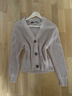 Beige stickad kofta från 157 - Säljer en beige stickad kofta från 157 i storlek S. Koftan har grovstickad struktur, djup v-ringning och tre stora knappar framtill. Modellen är lite croppad med ribbade muddar vid ärmslut och nederkant. Perfekt för lager-på-lager under kyliga dagar.